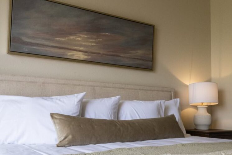 Close up view of cream upholstered headboard, white and tan pillows, white bedding, nightstand with lamp, and artwork above the bed