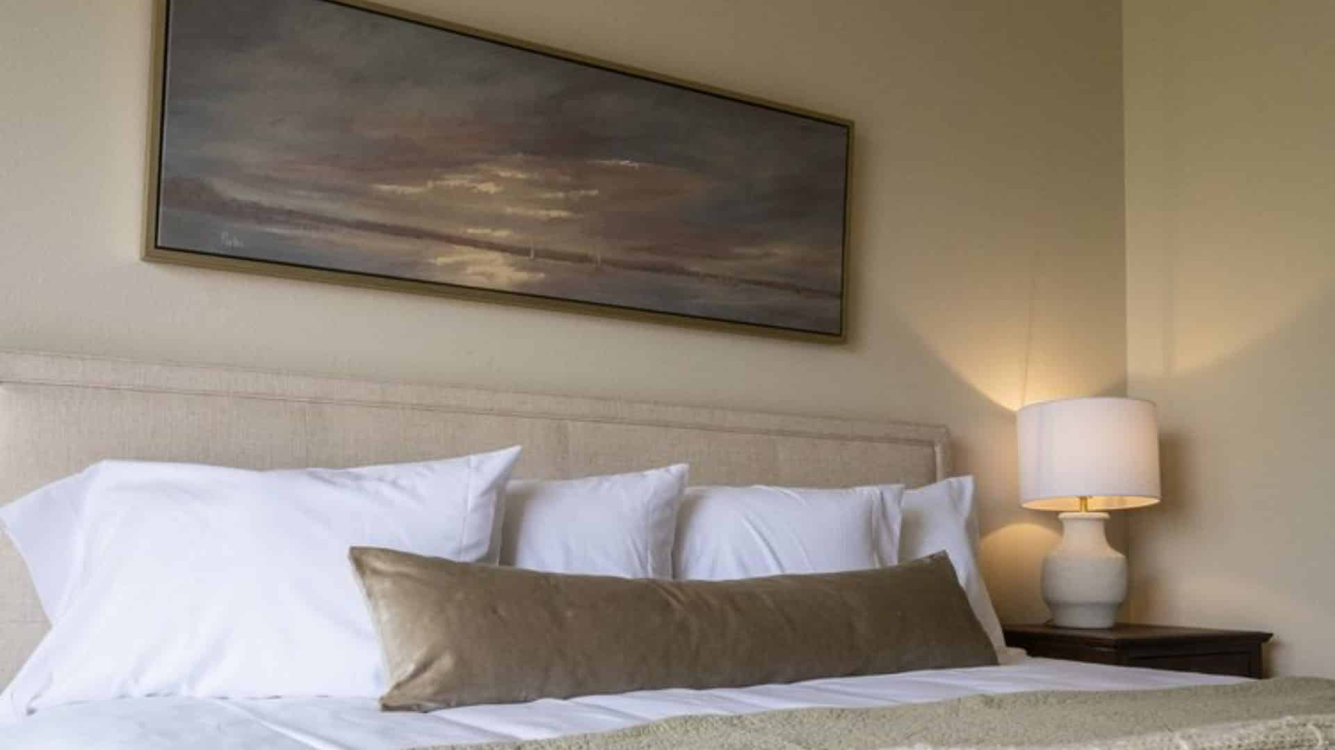 Close up view of cream upholstered headboard, white and tan pillows, white bedding, nightstand with lamp, and artwork above the bed